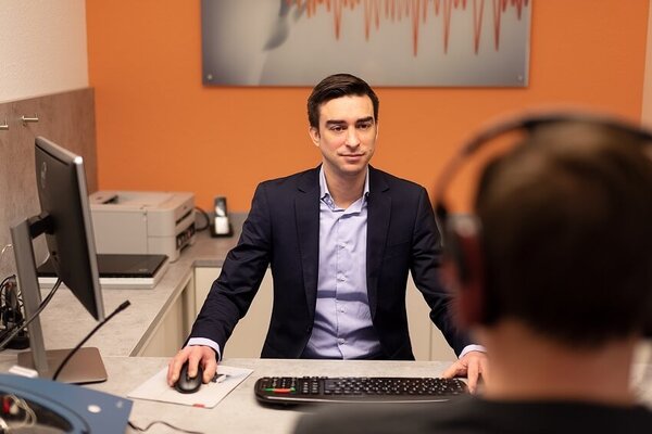 Mitarbeiter eines Hörakustikers berät Kunden am Schreibtisch in moderner Hörgeräte-Filiale.