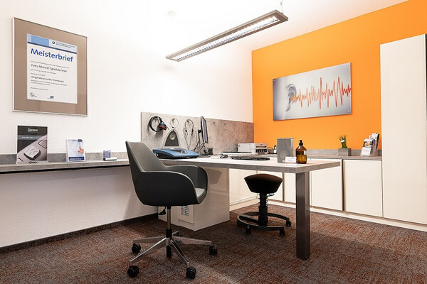Modern eingerichtetes Hörakustiker-Büro mit Schreibtisch, Audiogramm-Bild und Fachauszeichnungen.